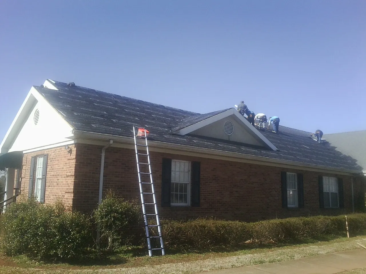 Crew tearing shingles off a brick ranch roof with a ladder set — Metal Roof Repair work by our crew in Ellenton